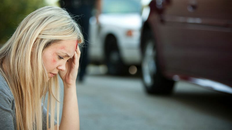 Woman holding her head sitting on the side of the road after a car accident