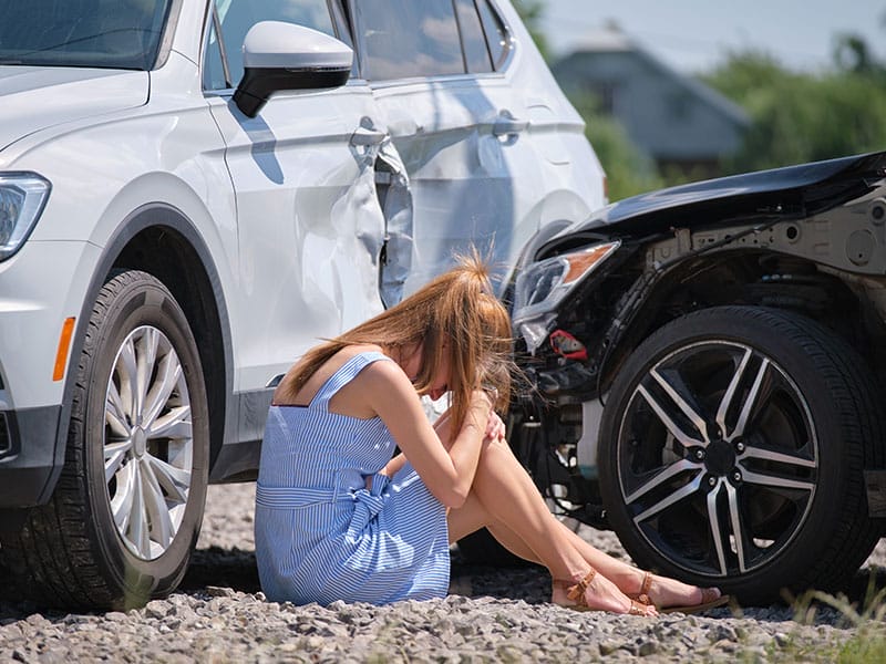 A woman sits on the ground next to her car after a car accident.