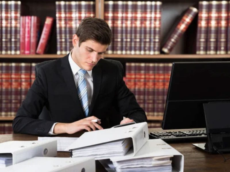 A man in a suit sits at a desk in front of a stack of books.