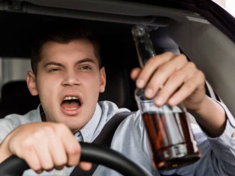 A man driving a car with a beer bottle in his hands.