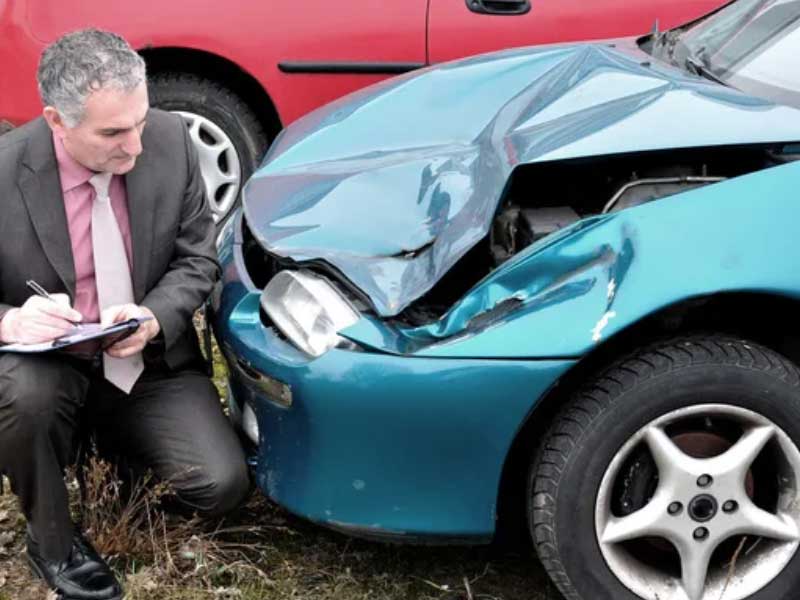 Hombre con traje inspeccionando y estimando el valor de la reclamación por accidente en un coche azul dañado.