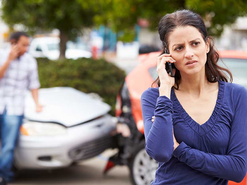 Mujer hablando por teléfono con expresión preocupada después de un accidente automovilístico mientras un hombre habla por teléfono en el fondo.