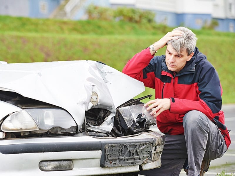 A distressed man examines his damaged white car with a visibly crumpled hood and broken headlights.