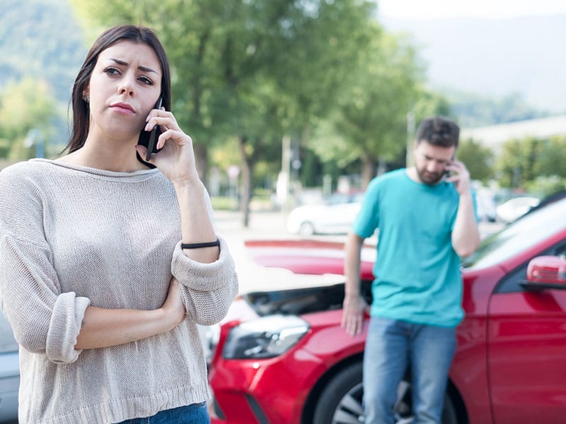 A woman talking on a phone looks worried beside a red car that has a minor collision with another car while a man stands behind with a confused expression.