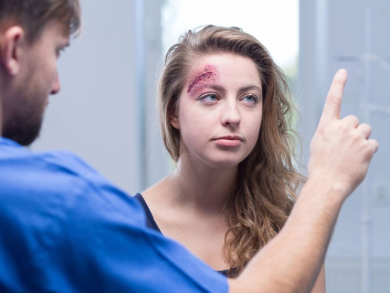 A medical professional is examining a woman with a visible injury on her forehead. The woman appears attentive as the professional holds up a finger for her to follow.