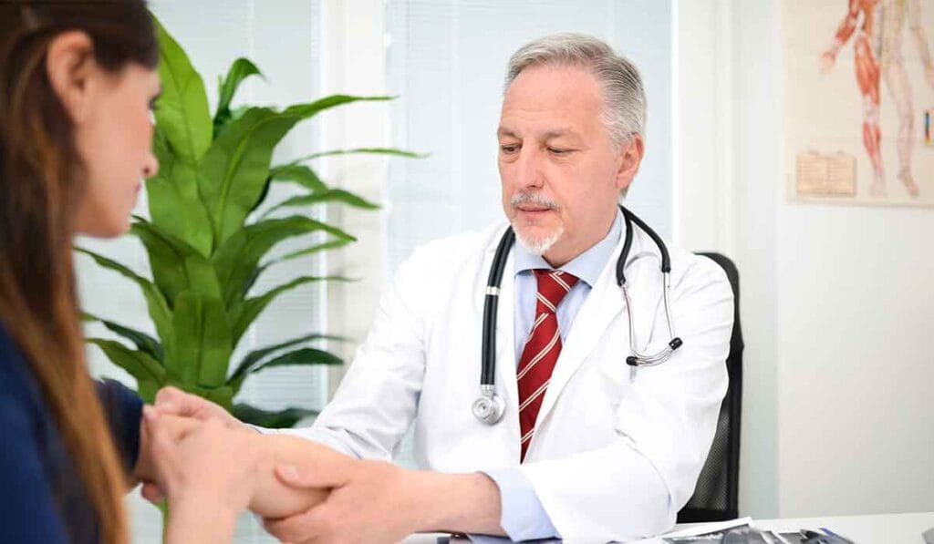 A doctor in a white coat examines a patient's arm in a medical office, with a plant and anatomical chart visible in the background.