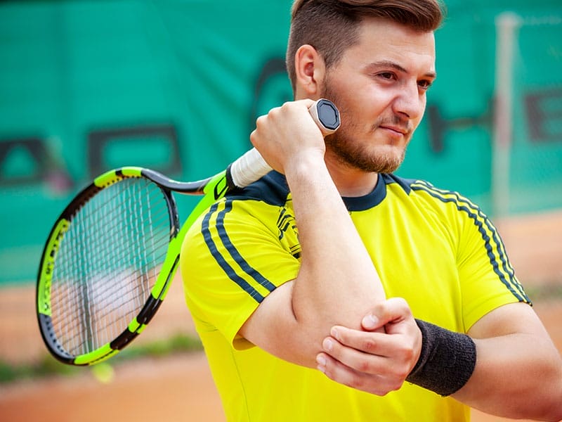 A man in a yellow shirt holds a tennis racket over his shoulder while stretching his arm on a tennis court.
