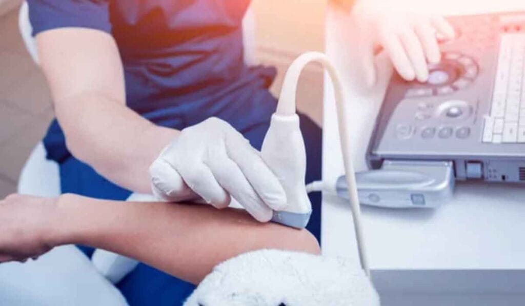 A medical professional performs an ultrasound on a patient's arm using a handheld device, while monitoring the results on a screen.