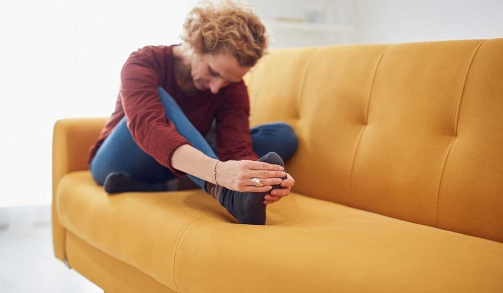 A person sitting on a yellow couch, stretching forward to touch their foot while wearing a red shirt and blue pants.