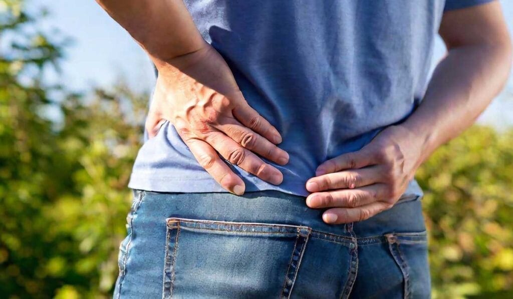 Una persona que viste jeans y una camisa azul se sostiene la espalda baja con ambas manos, de pie al aire libre.