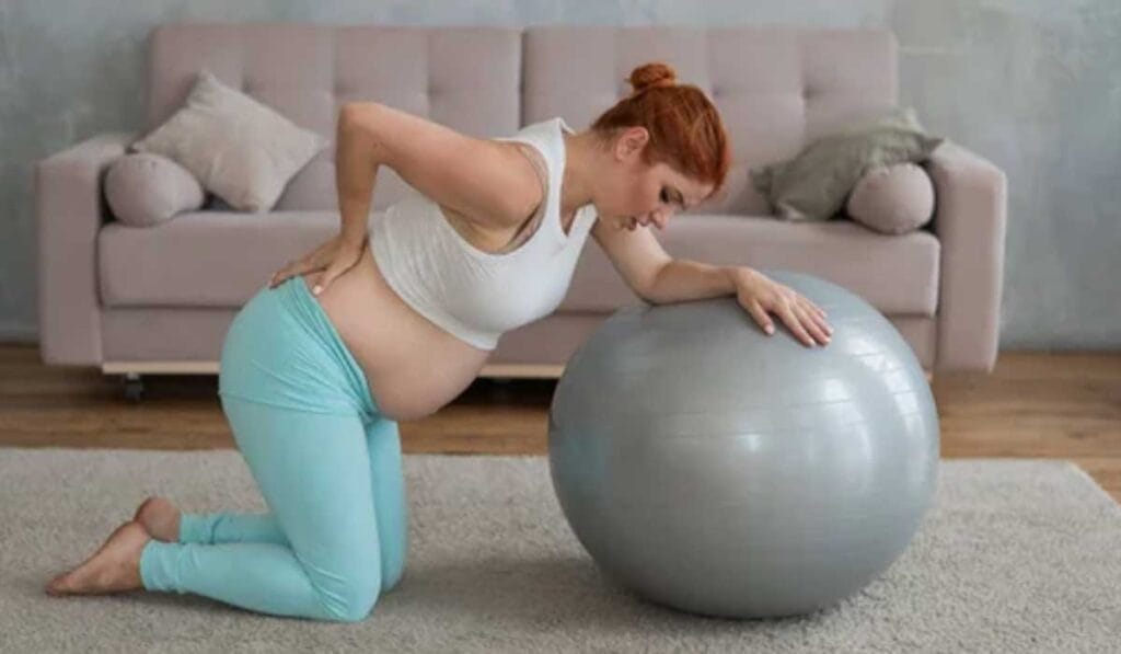 A pregnant woman kneels on a rug, supporting herself on a large exercise ball with one hand and holding her lower back with the other, in a living room with a sofa in the background.