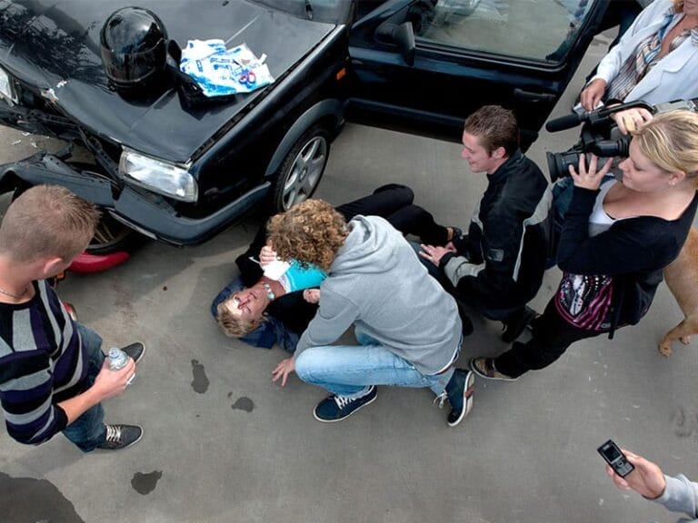 Person lying injured on the ground near a car with a damaged front, surrounded by people, one taking a video and another providing first aid.