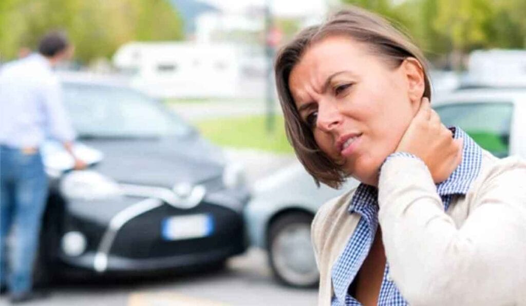 A woman with a pained expression touches her neck; blurred in the background, two men are inspecting damaged cars after an accident.