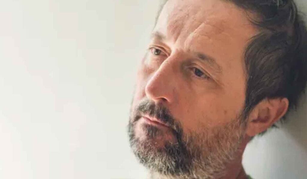 A man with short, dark hair and a beard leans his head against a white wall, looking to the side with a contemplative expression.