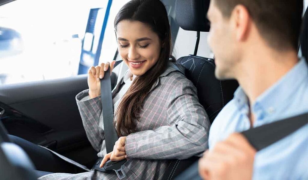 Two individuals sitting in a car, both fastening their seat belts. The person seated on the left is wearing a plaid jacket, and the person on the right is wearing a light blue shirt.