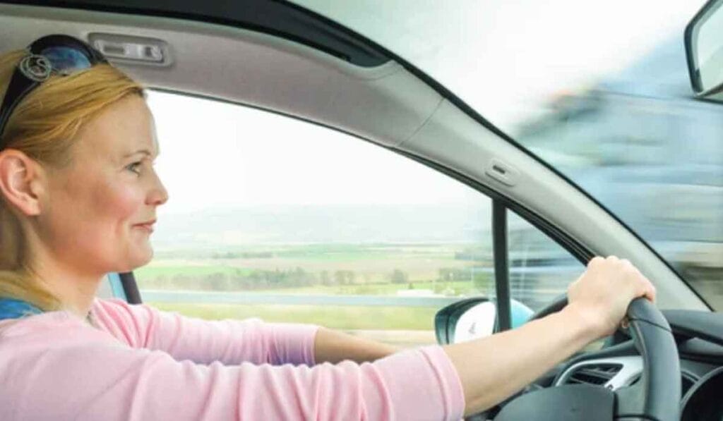 A woman with blonde hair is driving a car, focusing on the road ahead. The view through the window shows a blurred landscape due to the car's speed.