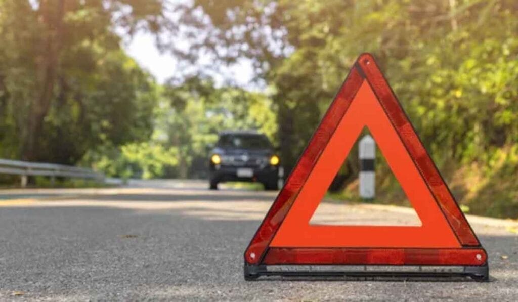 Close-up of a red triangular warning sign on a road with a blurred car approaching from the background under a canopy of trees.