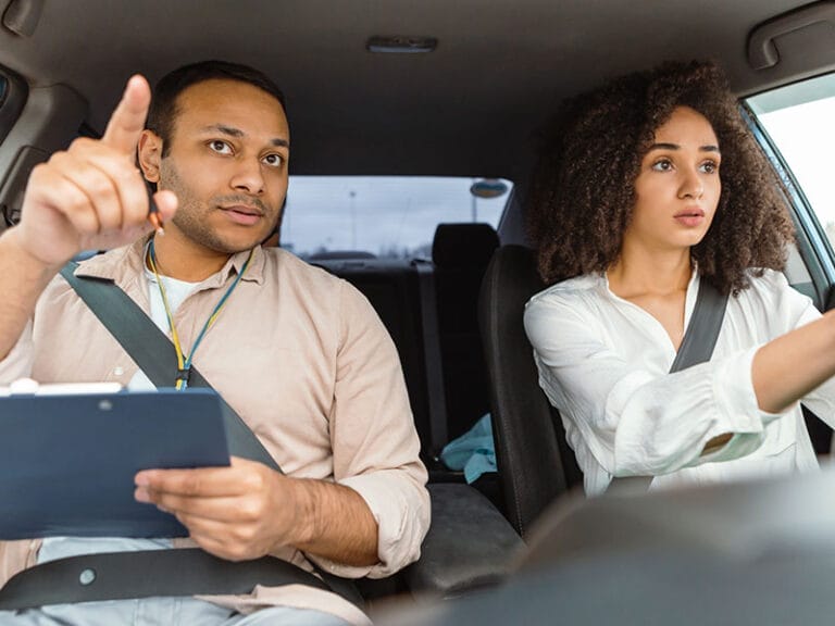 A man with a clipboard gestures while a woman drives a car, both wearing seatbelts.