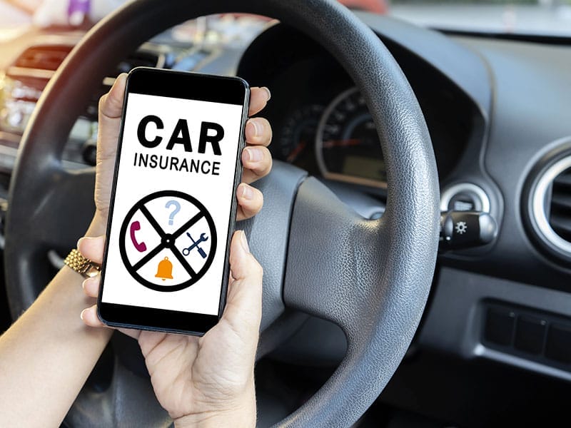 Person holding a smartphone with a car insurance app on the screen, sitting in the driver's seat with hands on the steering wheel.